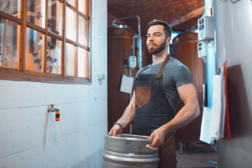 Wall Mural - A young brewer in an apron holds a barrel with beer in the hands of a brewery