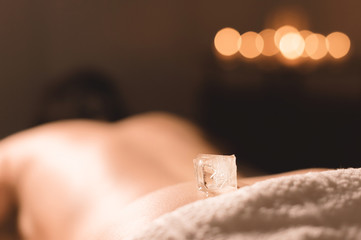 Wall Mural - Close-up of a transparent ice cube lying on the lower back of a young girl lying on a couch for spa treatments against the backdrop of a dark room and burning candles. Spa treatments. Small DOF