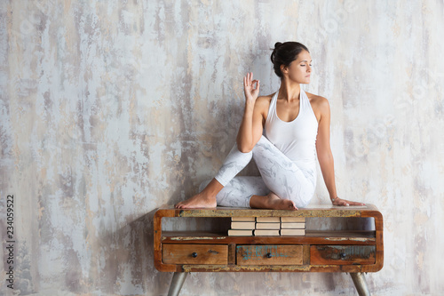 Fotografie, Obraz Slim beautiful young woman yoga instructor doing asana sitting on a vintage console against the background of a concrete wall