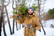 © artiemedvedev - Bearded man carrying freshly cut down christmas tree in forest. Lumberjack holds axe and fir tree on his shoulder in the woods. Irresponsible behavior towards nature, save forest, keep green concept.