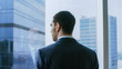 © Gorodenkoff - Medium Shot of the Thoughtful Businessman wearing a Suit Standing in His Office and Contemplating Next Big Business Deal, Looking out of the Window. Big City Business District Panoramic Window View.