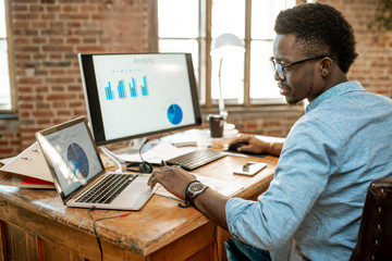 Wall Mural - young african man working with analitycs sitting at the beautiful working place with computer, lapto