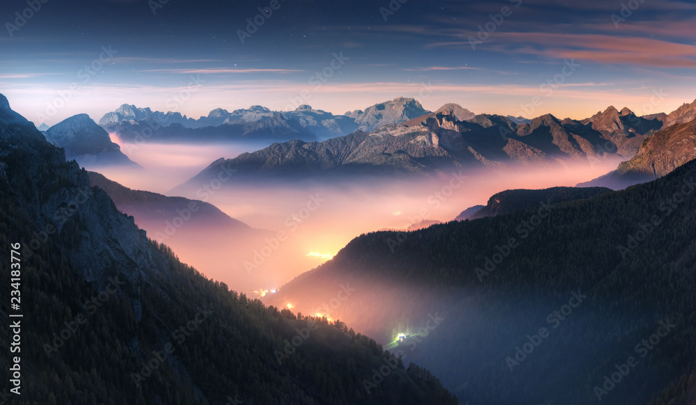 Mountains in fog at beautiful night in autumn in Dolomites, Italy. Landscape with alpine mountain valley, low clouds, forest, colorful sky with stars, city illumination at dusk. Aerial. Passo Giau - obrazy, fototapety, plakaty 