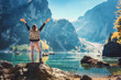 Standing woman on the stone with raised up arms on the coast of Braies lake at sunrise. Autumn in Dolomites, Italy. Landscape with happy girl, mountains, water with reflection, fall trees and blue sky