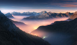 Fototapeta Fototapety góry  - Mountains in fog at beautiful night in autumn in Dolomites, Italy. Landscape with alpine mountain valley, low clouds, forest, colorful sky with stars, city illumination at dusk. Aerial. Passo Giau