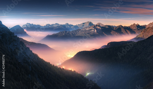 Fototapeta na wymiar Mountains in fog at beautiful night in autumn in Dolomites, Italy. Landscape with alpine mountain valley, low clouds, forest, colorful sky with stars, city illumination at dusk. Aerial. Passo Giau