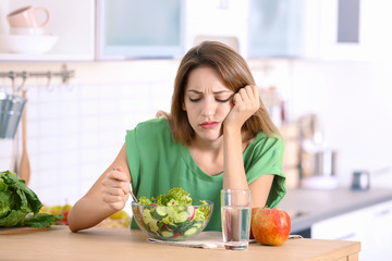 Wall Mural - Unhappy woman eating vegetable salad at table in kitchen. Healthy diet