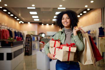 African american girl shopping gifts in mall on christmas sale. New year holidays concept. Smiling attractive mixed racial woman with colorful paper gift boxes wearing christmas hat in store or shop.