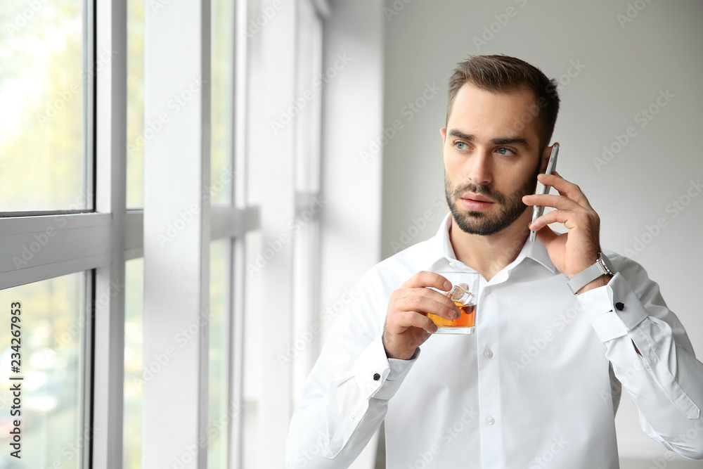 Young man with glass on whiskey talking by phone near window
