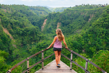 Wall Mural - Family vacation lifestyle. Young woman stand on edge of overhanging bridge on high cliff. Happy girl looking at stunning tropical jungle view. Tukad Melangit is popular travel destination in Bali.