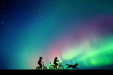 Poster - Children on bicycles and running dog in park at night. Vector illustration with silhouettes of boy and girl on bikes. Northern lights in starry sky. Colorful aurora borealis