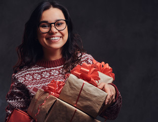 Portrait of a happy brunette girl wearing eyeglasses and warm sweater holding a gifts boxes, isolated on a dark textured background.