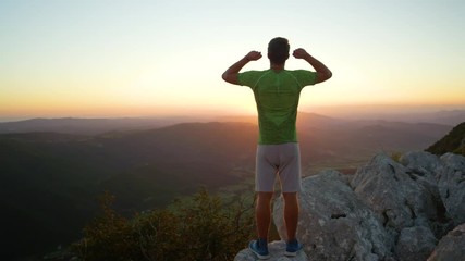 Wall Mural - SLOW MOTION, CLOSE UP, LENS FLARE, COPY SPACE: Male tourist celebrates reaching top of mountain at sunrise. Cinematic shot of unrecognizable male hiker observing nature with arms raised above his head