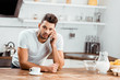 © LIGHTFIELD STUDIOS - young man leaning at kitchen table and talking by smartphone at morning