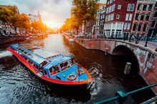 Tour Boat At Famous Dutch Canal On Sunset Evening. Traditional Dutch Bridges And Medieval Houses. Amsterdam Holland