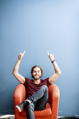 Wall Mural - Portrait of a young caucasian bearded man with long hair showing with hands on the colorful background sitting on the chair. Image with copy space