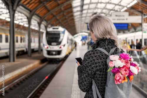 Fotografía Blonde woman with backpack with bouquet of flowers is holding smartphone and waiting train
