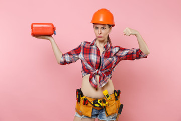 Strong young woman in orange helmet, plaid shirt, denim shorts, kit tools belt full of instruments, toolbox showing biceps, muscles isolated on pink background. Female male work. Renovation concept