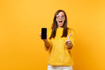 surprised young woman in heart glasses showing thumb up, holding mobile phone with blank black empty