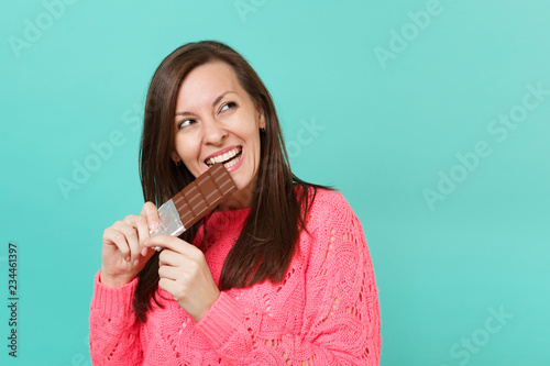 Fotografie, Tablou Pretty young woman in knitted pink sweater holding in hand biting chocolate bar looking aside isolated on blue turquoise wall background, studio portrait
