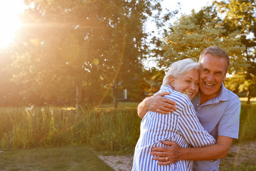 Portrait Of Loving Senior Couple Hugging Outdoors In Summer Park Against Flaring Sun