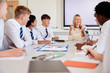 © Monkey Business - Female High School Teacher Sitting At Table With Teenage Pupils Wearing Uniform Teaching Lesson