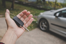 Close Up Of Man's Palm Holding Car Key On A Parked Car Background. Driver Holds The Vehicular Key On The Way To The Vehicle. Man Invites To Have A Drive In His Car. Expensive Present. Motor Purchase.
