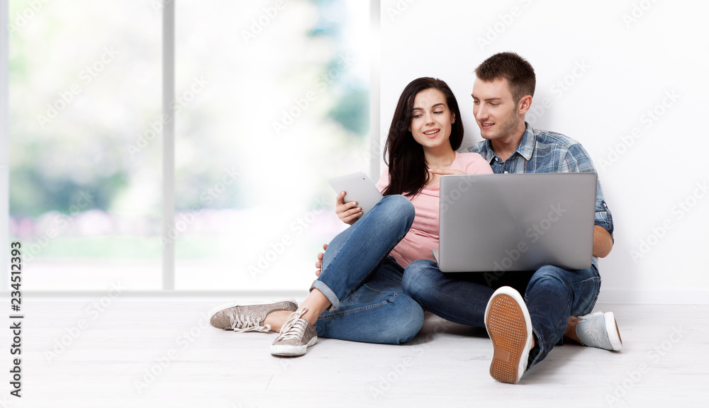 Happy young couple sitting on the floor with a laptop looking for their new home and furniture. Mock up for design.