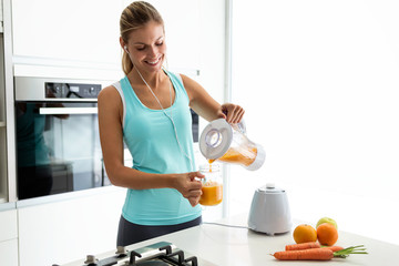 Wall Mural - Beautiful young sporty woman serving detox orange juice into glass while listening music in the kitchen.