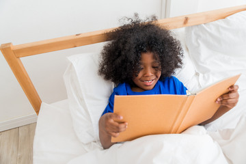 A cute little child African American girl reading a book while lying on the bed. Kid enjoy learning with happiness at home. Clever and smart learning concept