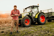 Male Farmer Working On Field Using Smartphone In Modern Agriculture - Tractor Background