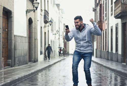 Fotografía Excited young man looking at cellphone with victorious expression in the middle of the street
