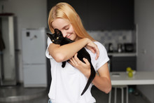 Close Up Of Young Blonde Girl At The Kitchen Holds On Hands, Caresses, Stroking, Hugs Black Little Cat, Cute Tenderly Smiling, Eyes Closed, Dressed In A Domestic White T-shirt, Kitchen Background