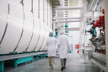 Wall Mural - Back view full length portrait of two factory workers wearing lab coats crossing production workshop, copy space