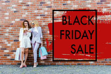 shocked young women with shopping bags taking selfie while standing in front of brick wall, black friday sale banner