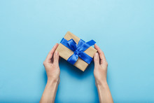 Female Hands Holding A Gift With A Blue Ribbon On A Blue Background. Concept Of A Gift For The Holidays, Birthday, Christmas, Wedding. Flat Lay, Top View