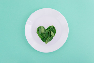 Wall Mural - Green leaves heart shape on white plate on green pastel background.