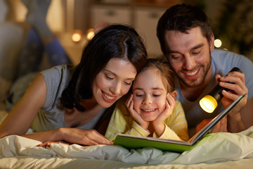 Poster - people and family concept - happy mother, father and little daughter reading book with torch light in bed at night at home