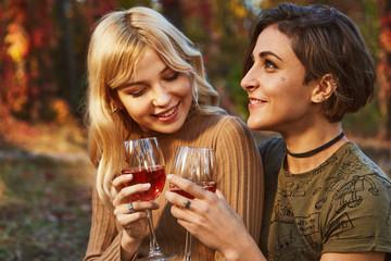 A couple of lesbian ladies in the park. The young pretty women holding glasses with red wine, smiling. Close up portrait of the blonde and brunette girls posing against the blurred nature background.