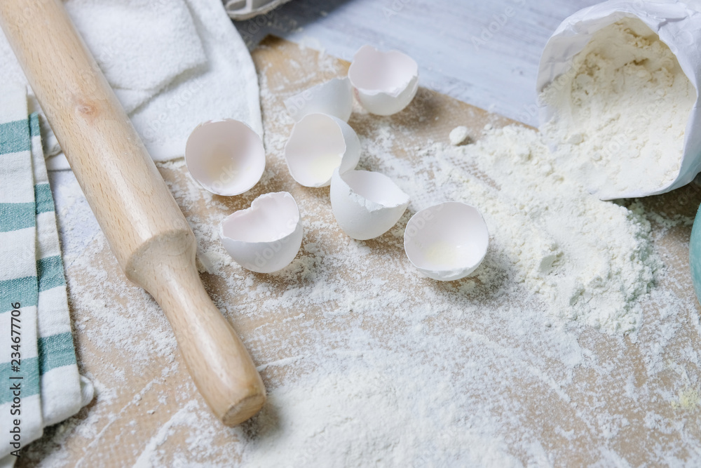 Table with ingredients for dough, raw eggs, flour, plate, napkins, rolling pin. Process of preparing pie, kitchen objects, flat lay with light white and blue colors space for text - obrazy, fototapety, plakaty 