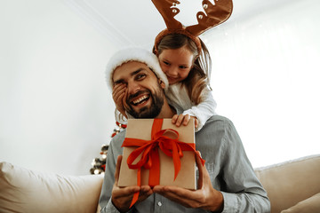 Christmas. Family. Love. Little girl is covering her dad's eyes making a surprise with a gift box. Near the Christmas tree at home