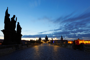 Wall Mural - Scenery of Charles Bridge before sunrise with street lamp lights glowing in blue twilight & silhouettes of majestic statues & towers of historical buildings under dawning sky in Old Town Prague, Czech