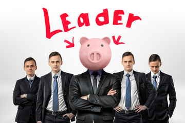 Several identical businessmen stand clothe to each other around one man with a piggy bank head under the Leader word. T