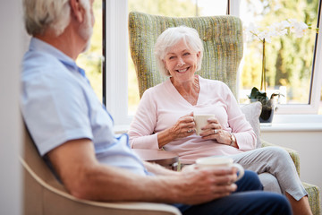 Senior Couple Sitting In Chairs And Chatting In Retirement Home