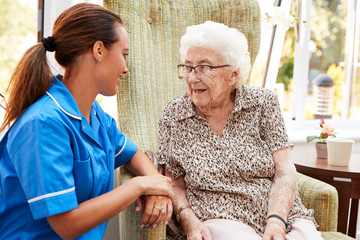 Senior Woman Sitting In Chair And Talking With Nurse In Retirement Home