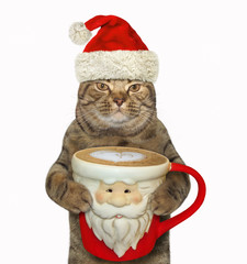 The cat in Santa Claus hat holds a big cup of black coffee. White background.