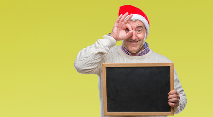 Canvas Print - Handsome senior man wearing christmas hat and holding blackboard over isolated background with happy face smiling doing ok sign with hand on eye looking through fingers