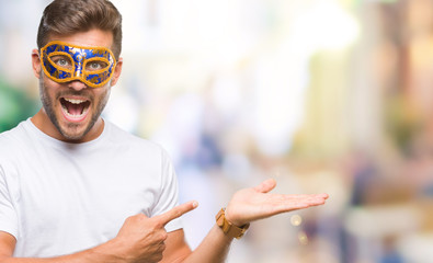 Sticker - Young handsome man wearing carnival mask over isolated background amazed and smiling to the camera while presenting with hand and pointing with finger.