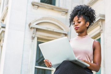 Way to Success. Young African American woman with afro hairstyle wearing sleeveless light color top, sitting by vintage office building in New York, looking down, reading, working on laptop computer..