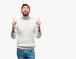 Adult hispanic man wearing winter sweater over isolated background amazed and surprised looking up and pointing with fingers and raised arms.